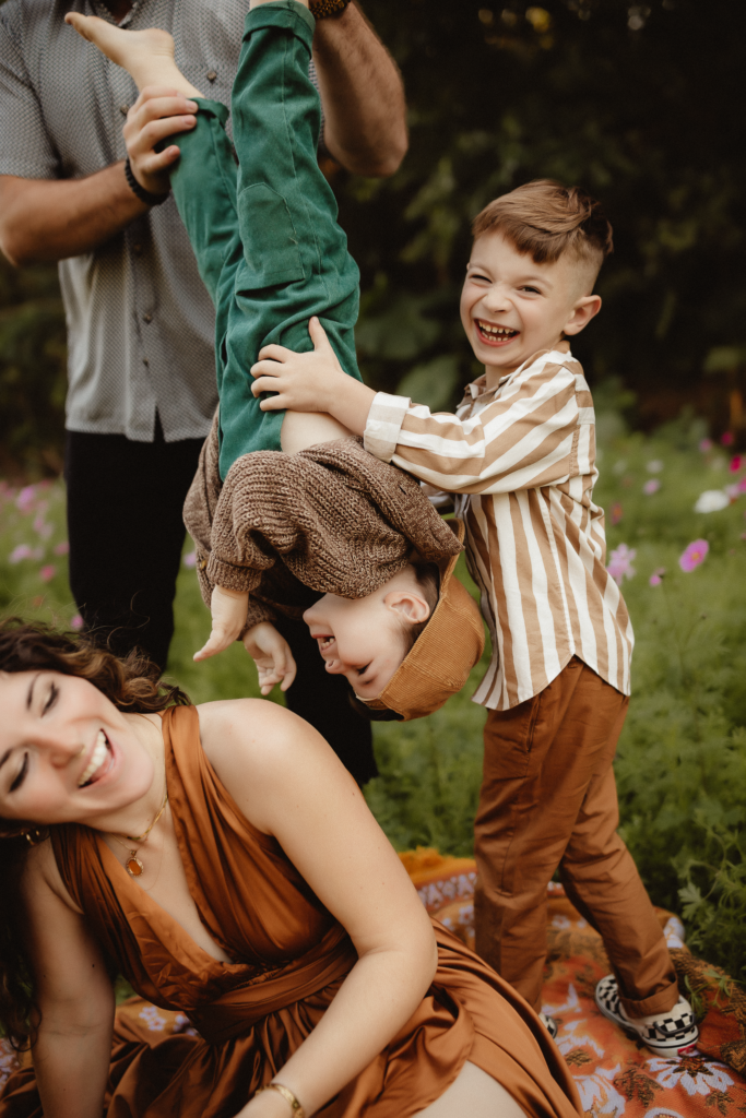 Dad playfully holds his son upside down in a flower field, eliciting laughter from both mom and brother, creating a joyful family scene, photographed by a washington family photographer