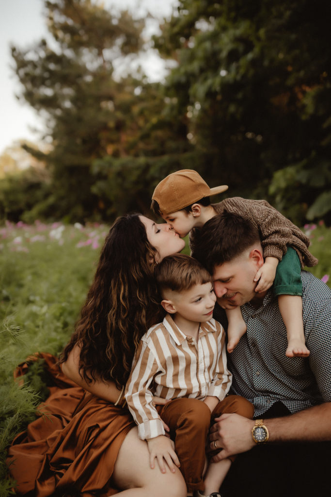 In a picturesque flower field, a son enjoys the view from his dad's shoulders, while mom sweetly kisses him. The whole family shares a serene moment, with dad's nose resting gently on his sons's temple being photographed by a washington family photographer