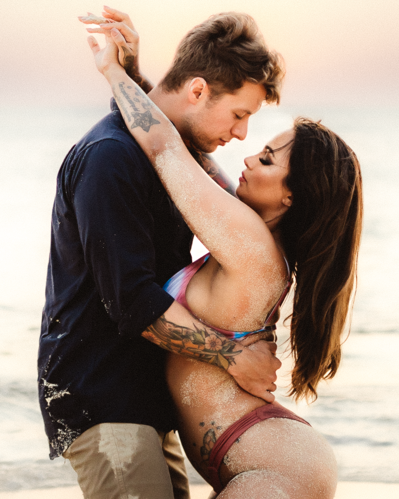 amidst the picturesque washington beach, a couple shares an intimate moment on their knees. The wife embraces the husband, her arms around his shoulders, while he tenderly holds her waist. Their eyes lock, creating a beautiful connection against the backdrop of the tranquil coastal scene being photographed by a washington couples photographer