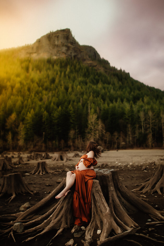a pregnant mom sitting on a stump while letting the wind take her hair, while rubbing on her belly and being photographed by a washington state maternity photographer