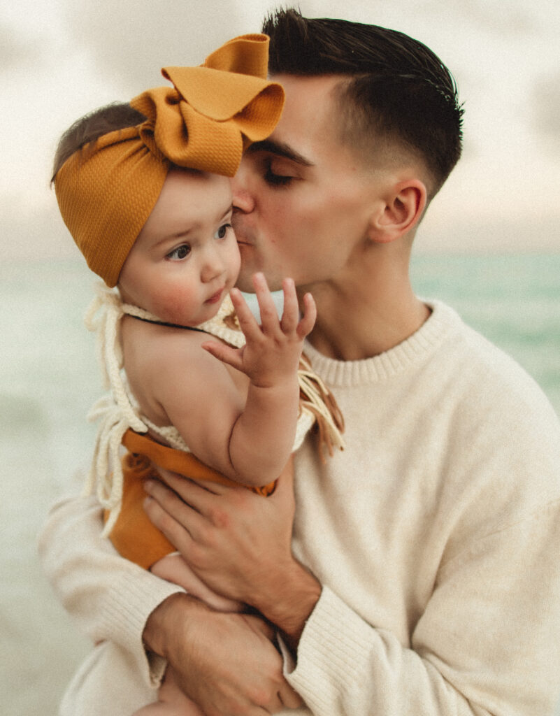 dad is holding his daughter at the beach in washington state while kissing on her cheek and being photographed by a washington state family photographer